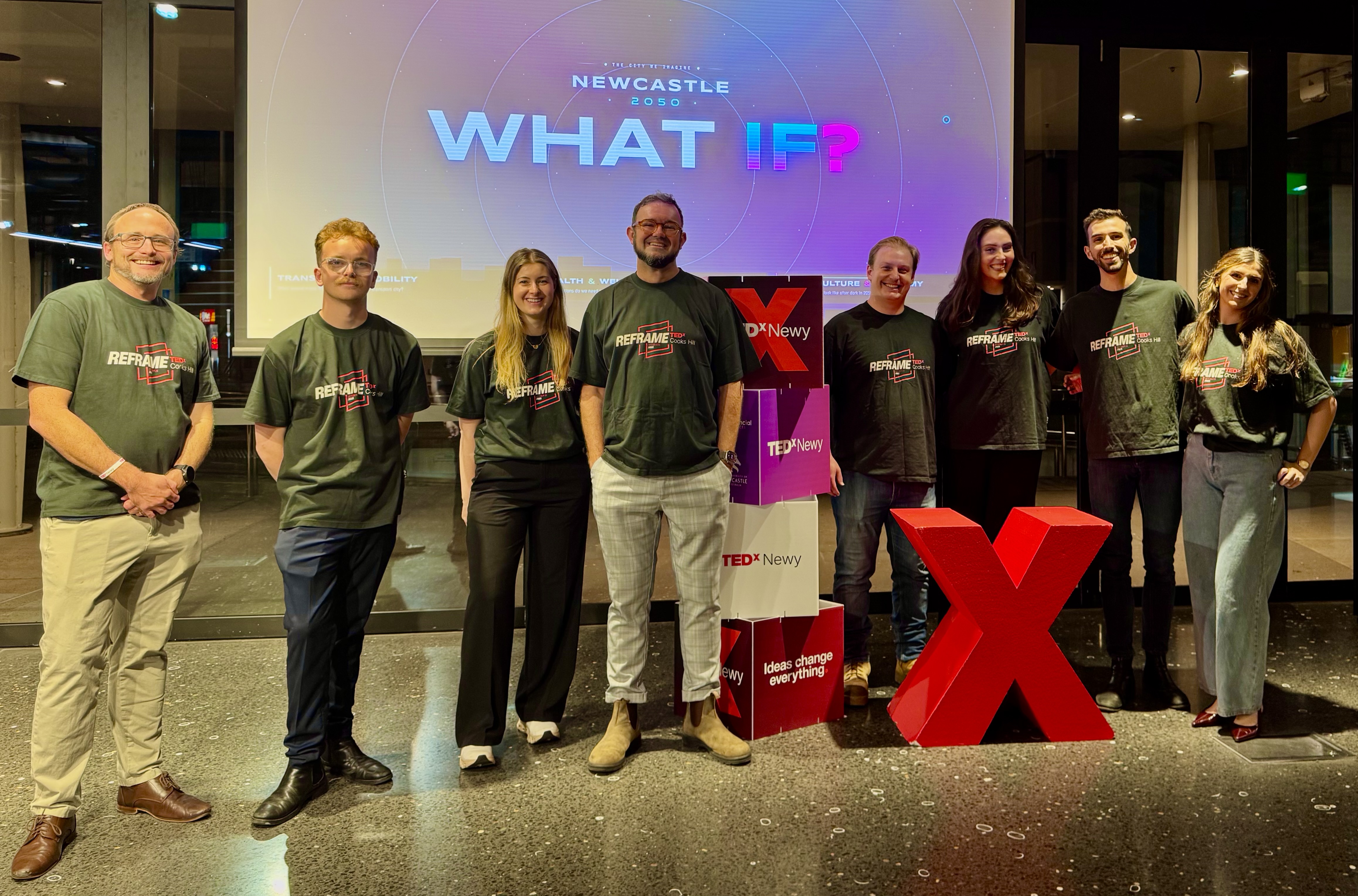 The TEDxNewy team standing in front of a 'What If?' projection and a large red X at the Q Building, University of Newcastle.